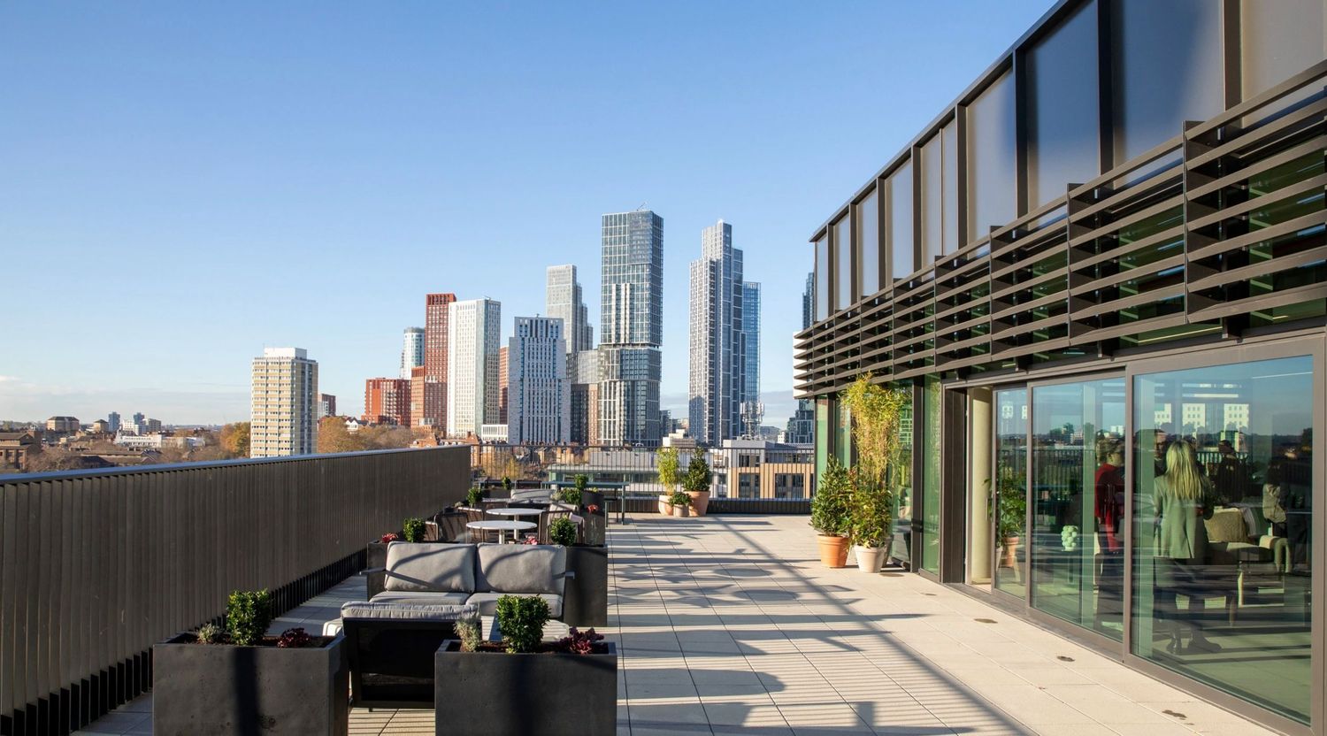 Rooftop terrace with city skyline views, modern architecture, potted plants, and glass building facade on a sunny day.
