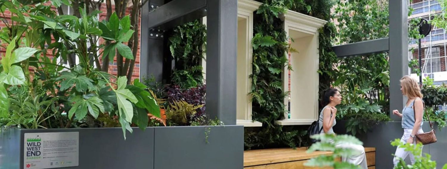 Two women walk past a modern green wall installation featuring lush tropical plants and white architectural frames in an urban setting.