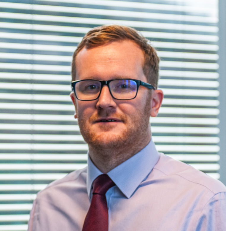 Professional man wearing glasses and a red tie, posing in a modern office setting with blinds in the background.