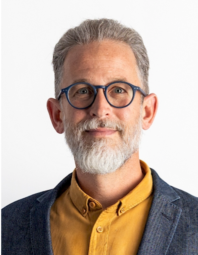 Middle-aged man with gray beard and round blue glasses wearing a yellow shirt and navy blazer against white background.