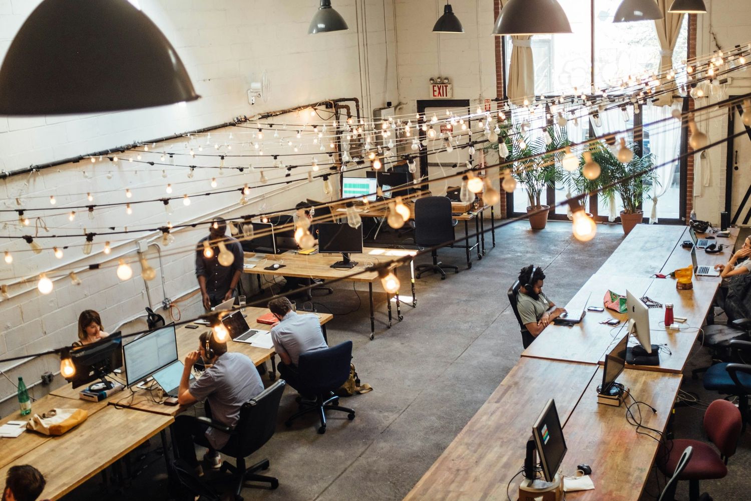 Modern open-plan coworking office space with string lights, exposed brick walls, and professionals working at wooden desks.