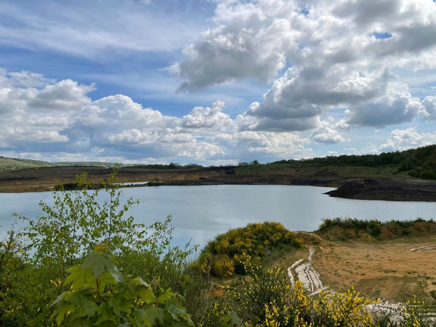 Scenic view of a tranquil lake surrounded by sandy shores and lush greenery under a cloudy blue sky.