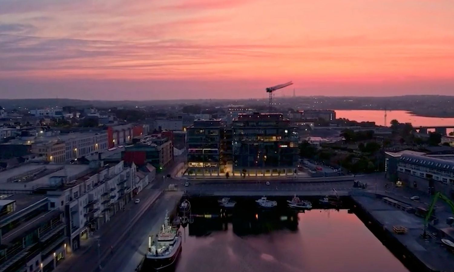 Aerial view of a coastal city at sunset with vibrant pink and orange sky reflecting over calm harbor waters.
