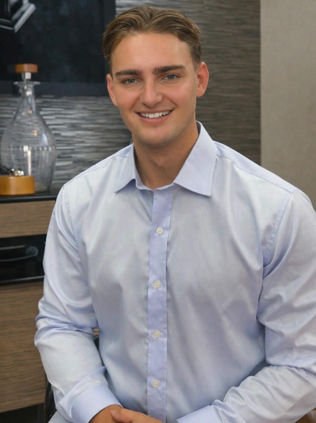 Smiling young professional man in a light blue dress shirt seated in a modern office interior.