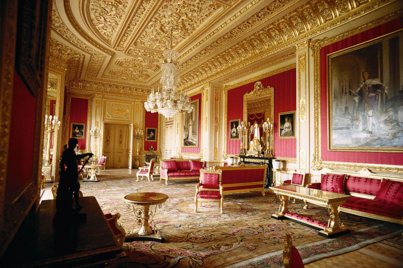 Opulent royal palace room with crimson walls, gold ornate ceiling, crystal chandelier, and elegant red velvet furniture.