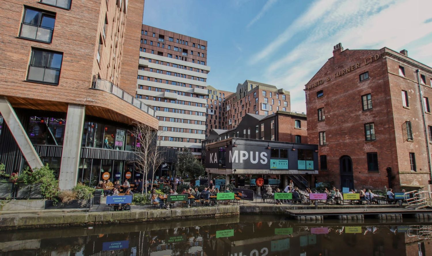 Vibrant canal-side scene at Manchester's Castlefield with historic brick buildings, modern architecture, and outdoor dining crowds.