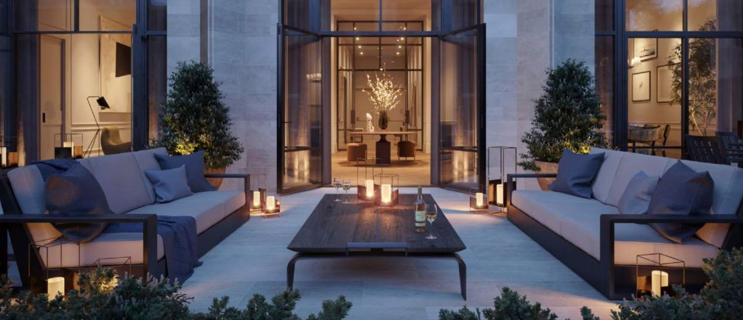 Elegant outdoor terrace with plush gray sofas, coffee table, lanterns, and lush greenery illuminated at dusk.