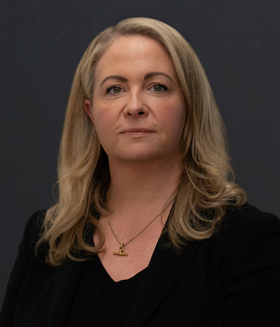 Professional headshot of a blonde woman in a black blazer wearing a gold necklace against a gray background.