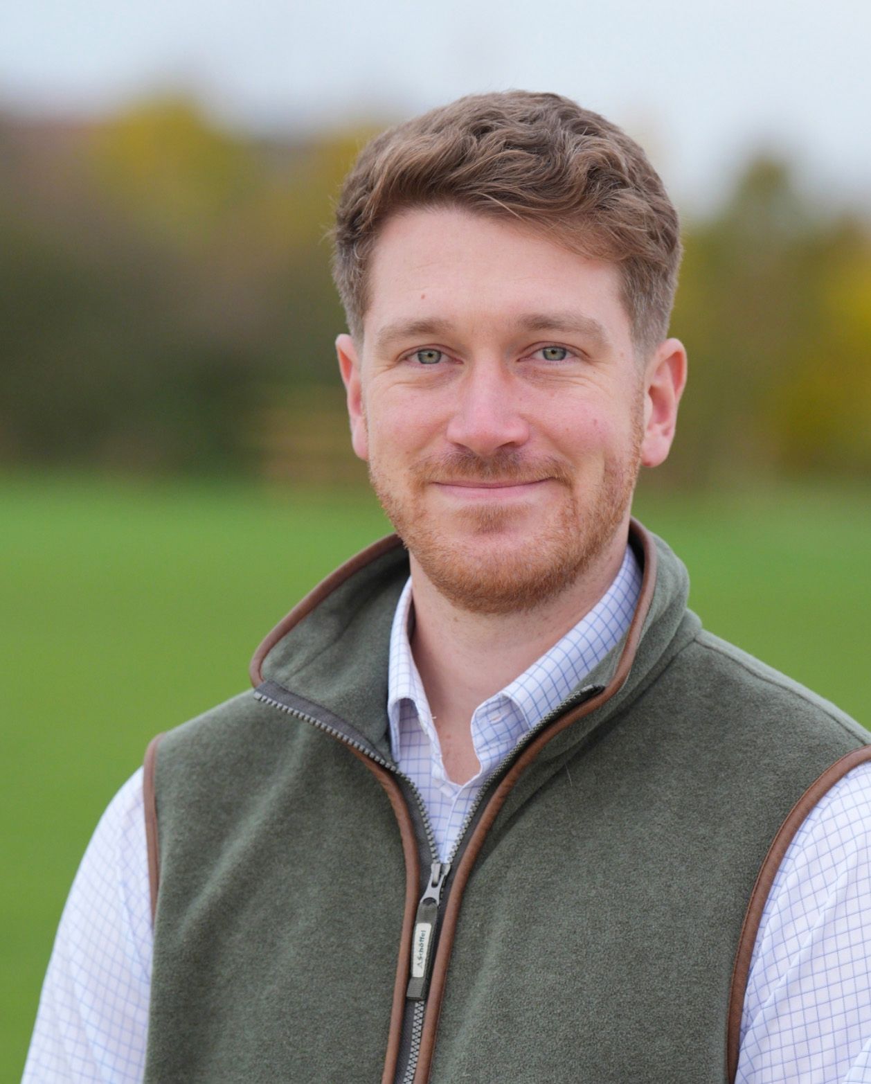 Smiling man with red beard wearing a green vest over a plaid shirt, standing outdoors in a green field.