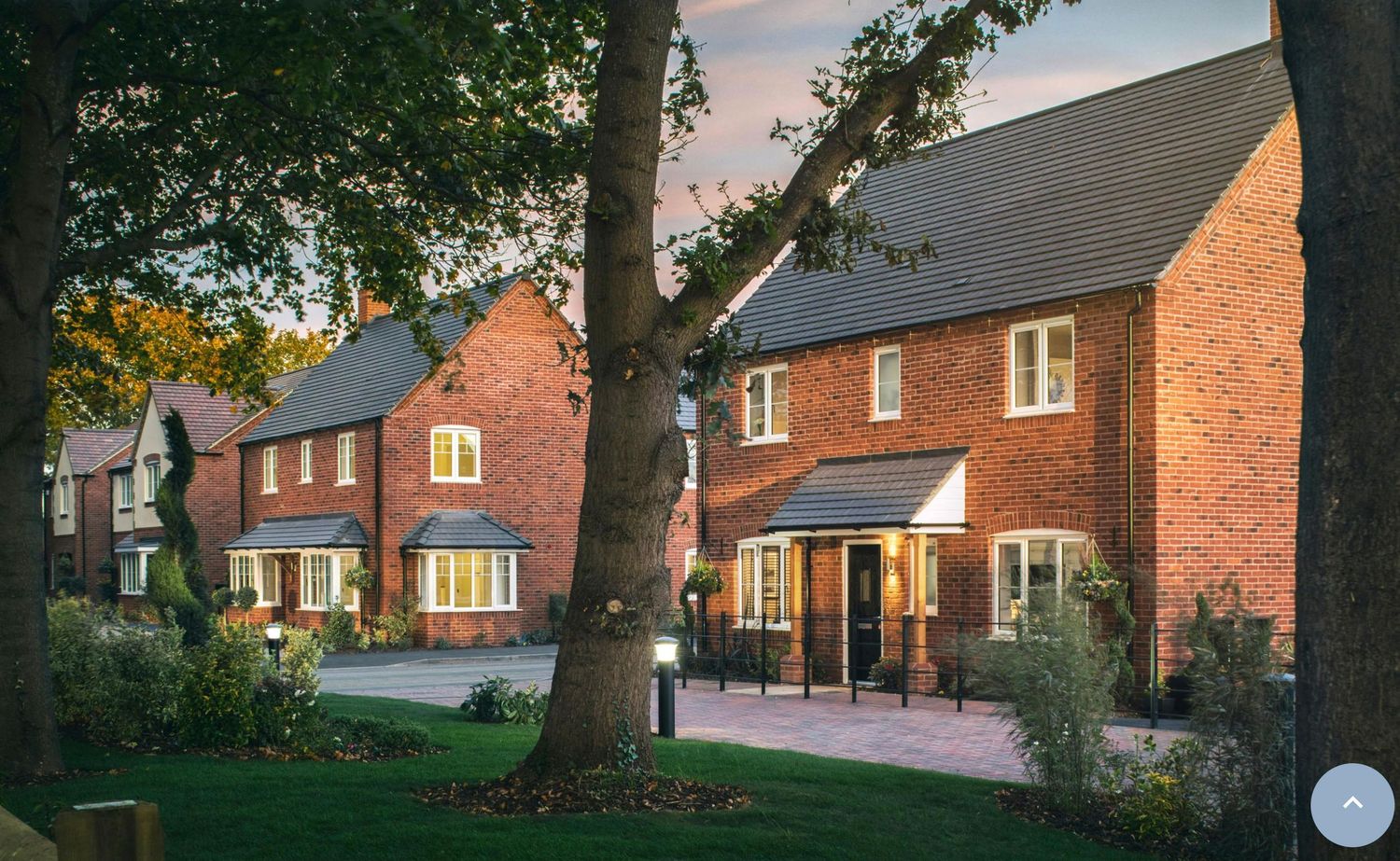 Modern red brick detached houses with manicured lawns and mature trees in a new residential development at dusk.