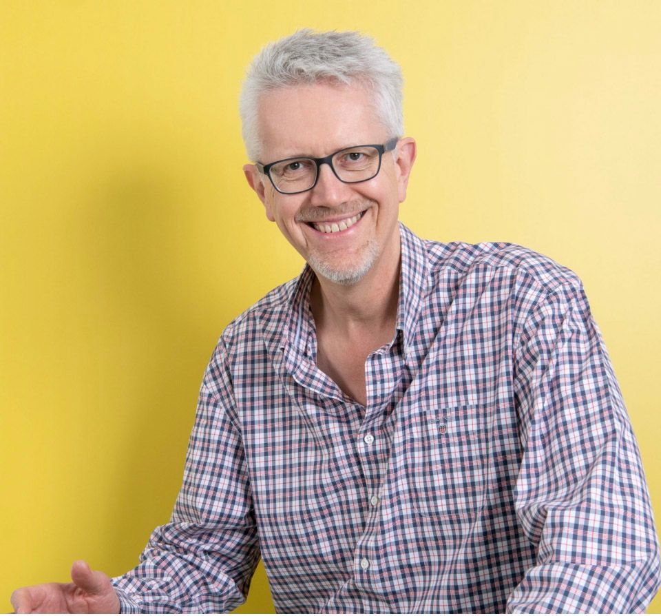 Smiling middle-aged man with gray hair and glasses wearing a plaid shirt against a yellow background.