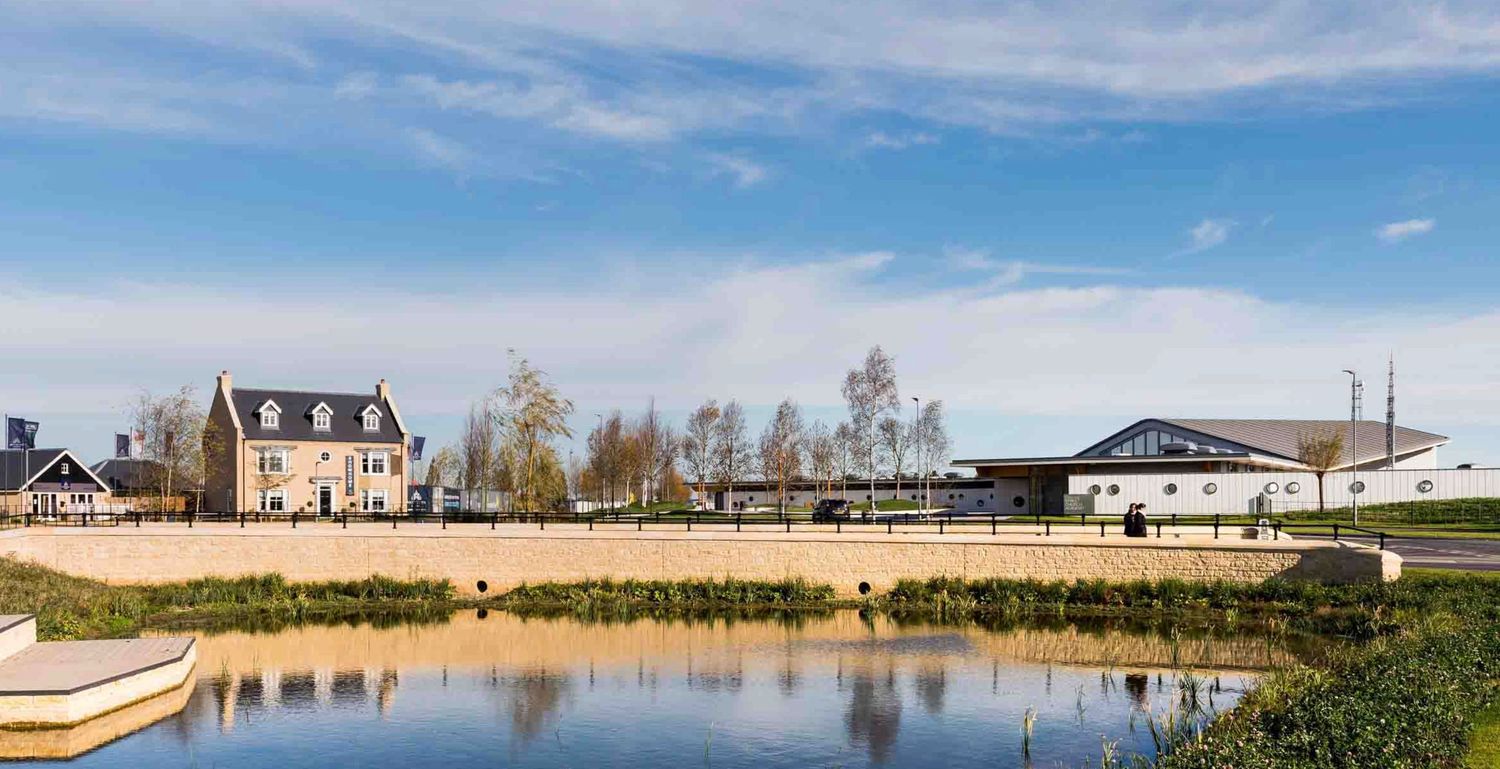 Scenic waterfront development with a historic house, modern building, and calm reflective pond under a blue sky.
