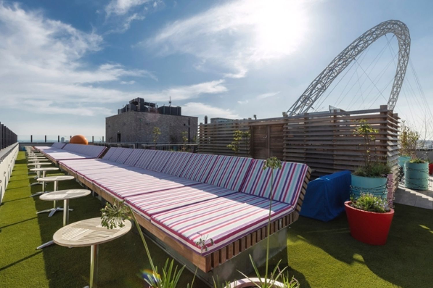 Rooftop terrace with striped loungers on artificial grass, white side tables, and Wembley arch visible under sunny blue sky.