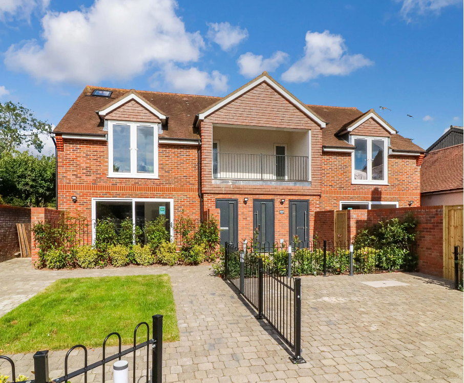 Modern red brick semi-detached house with balcony, black iron gates, block paved driveway, and manicured garden under blue sky.