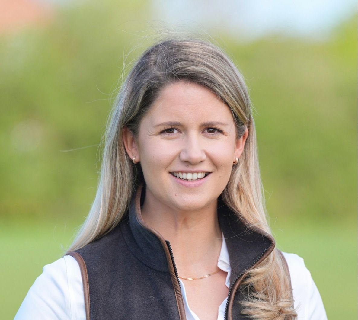 Smiling woman with long blonde hair wearing a dark vest over white shirt outdoors in a green field.