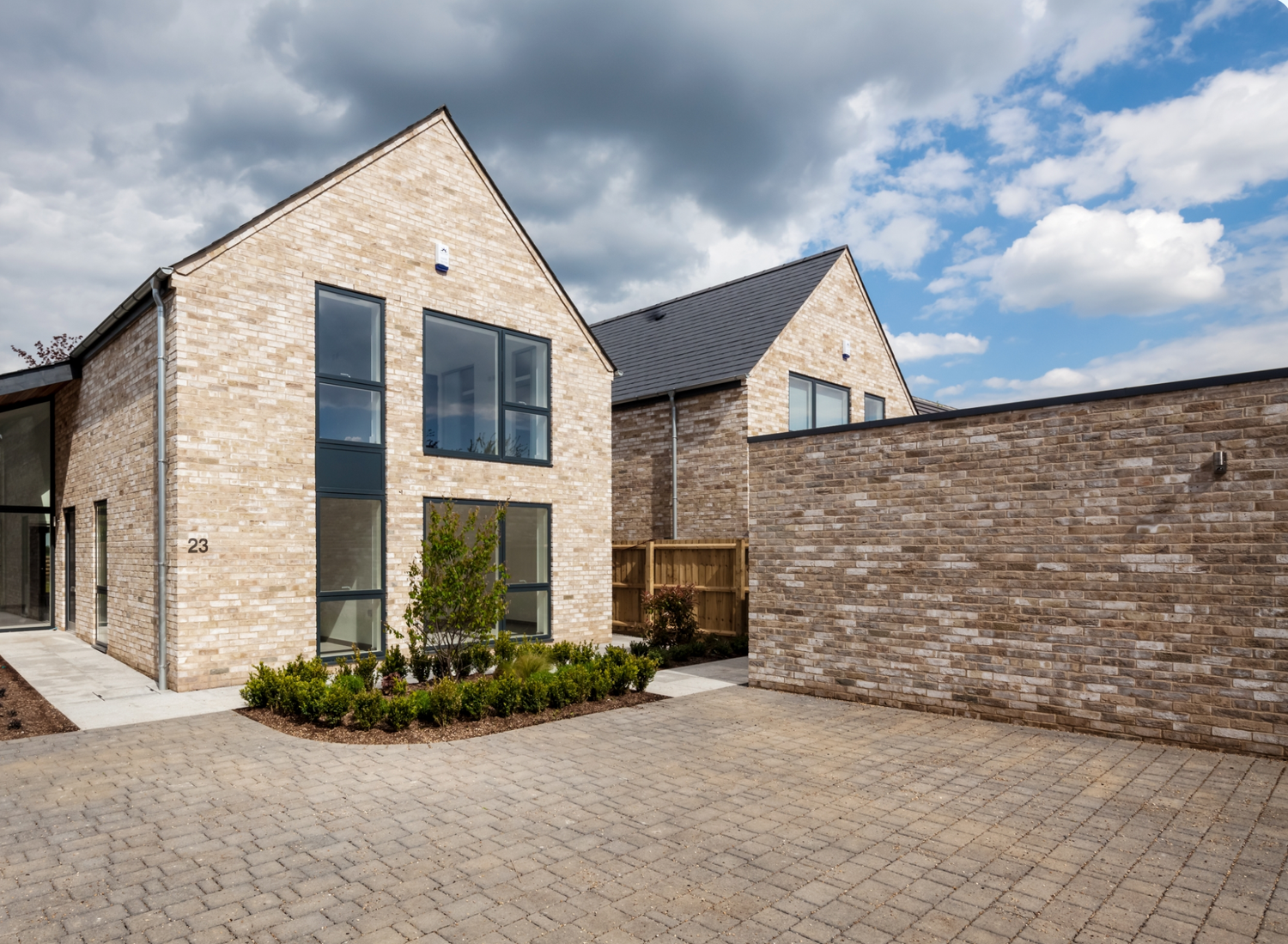 Modern stone brick house with large windows, black roof, and paved driveway under a cloudy sky.