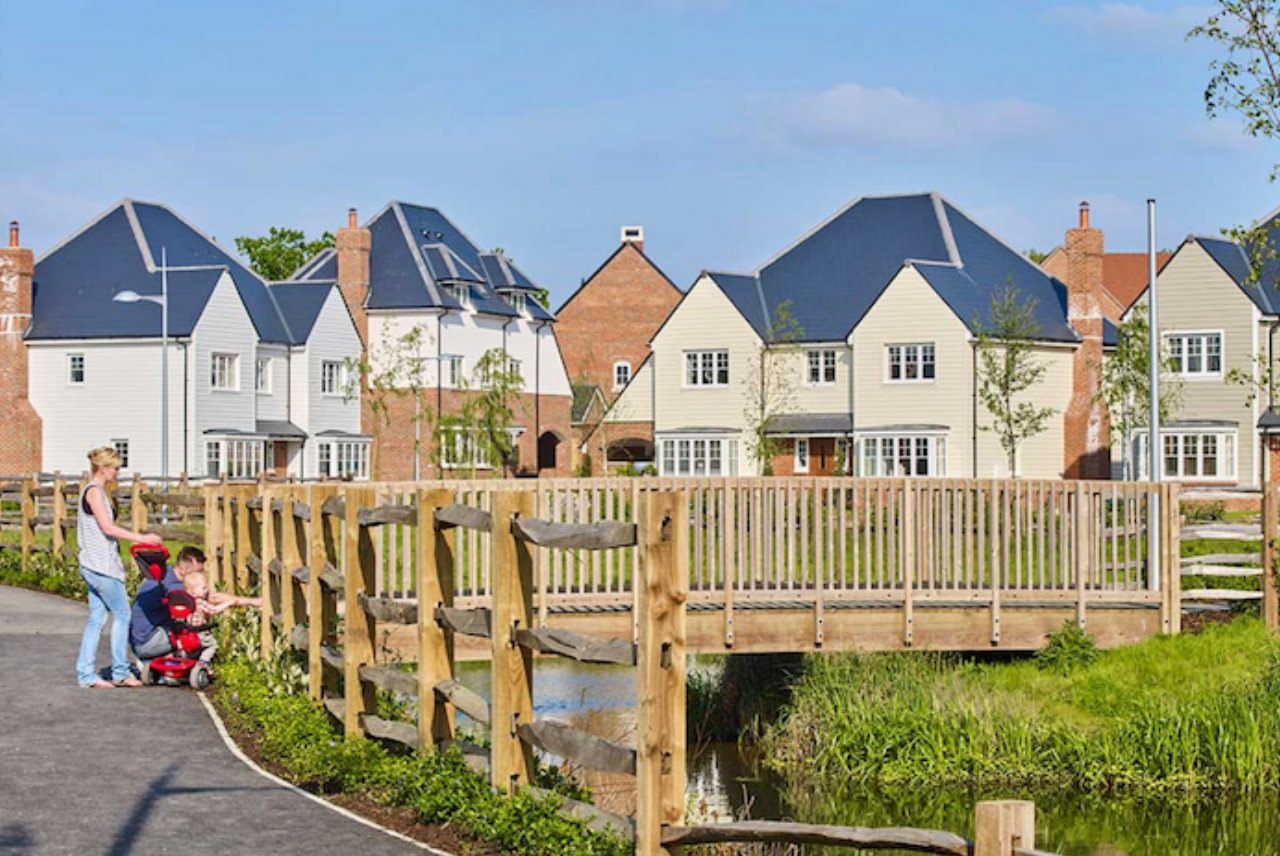 Woman with stroller on wooden bridge in modern UK housing development with white and brick homes.