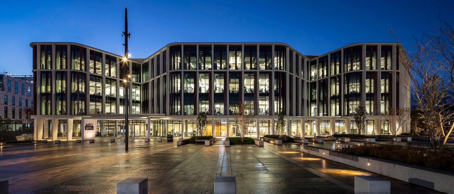Modern illuminated office building with curved facade and large windows photographed at dusk in an urban plaza.