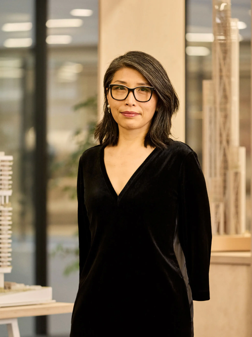 Woman in black velvet dress and glasses stands confidently in a modern office with architectural models.