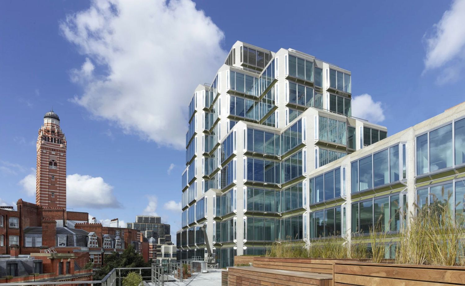 Modern glass office building with rooftop terrace beside historic red brick clock tower under blue sky.