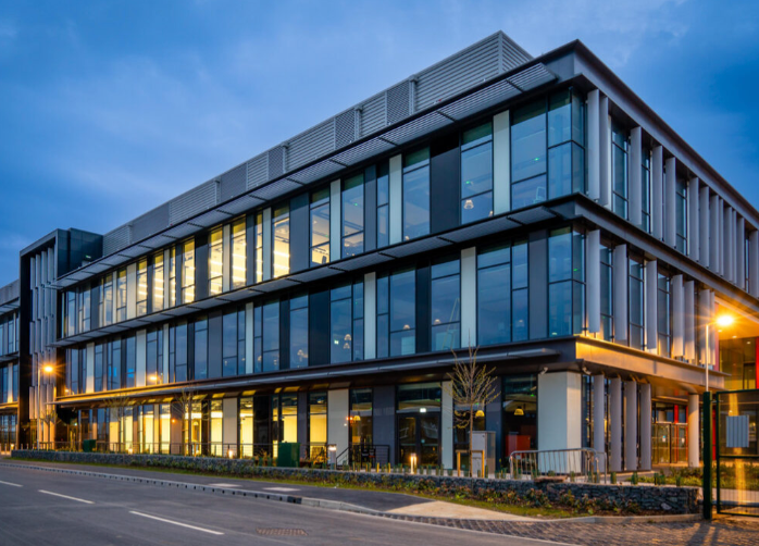 Modern multi-story glass office building illuminated at dusk with warm interior lighting on a quiet urban street.