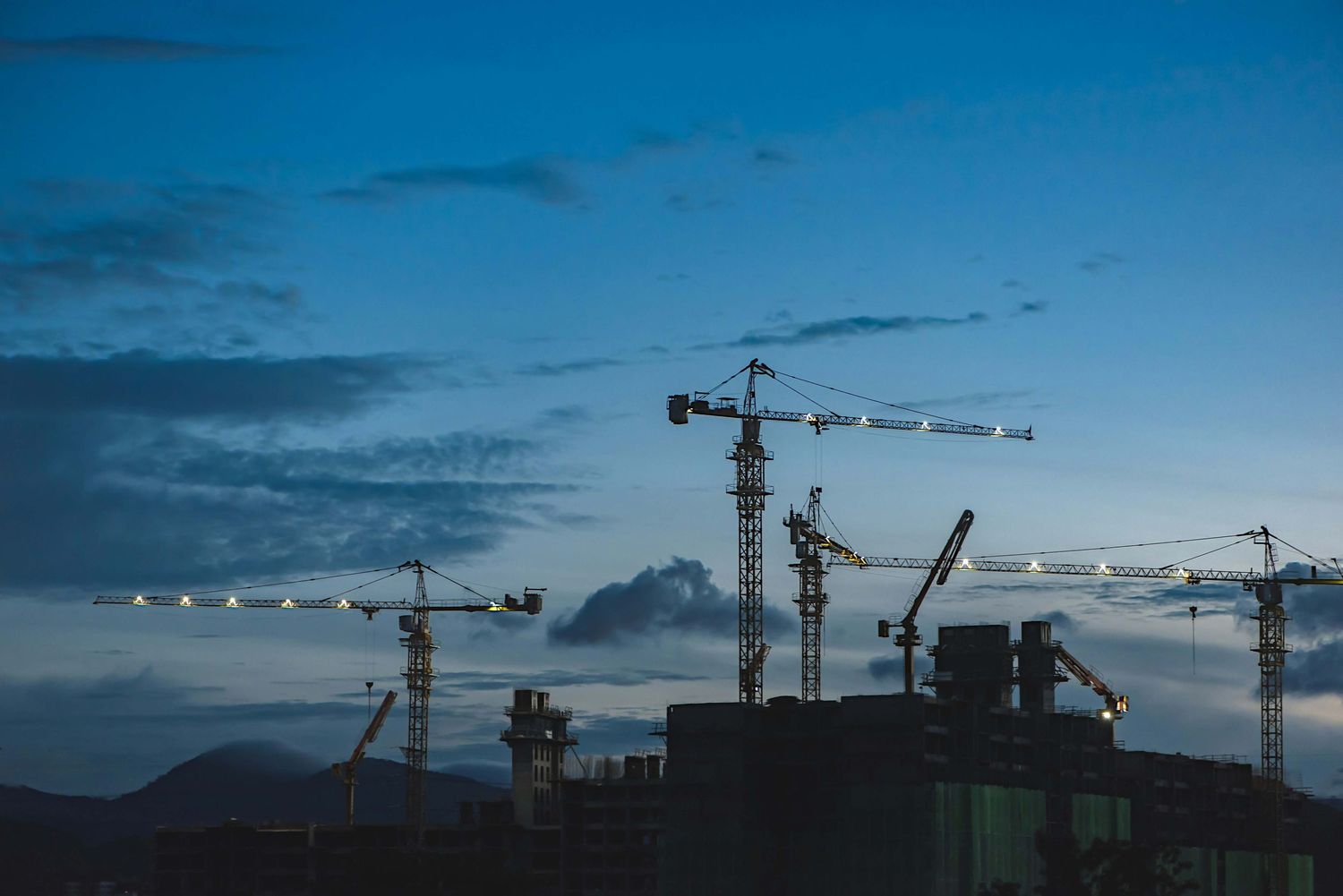 Multiple construction cranes silhouetted against a dramatic blue dusk sky at an active building site.