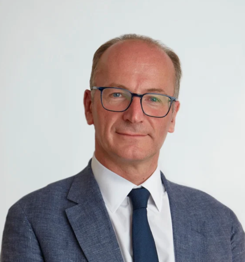 Professional headshot of a middle-aged man wearing glasses, blue tie, and gray suit jacket against white background.