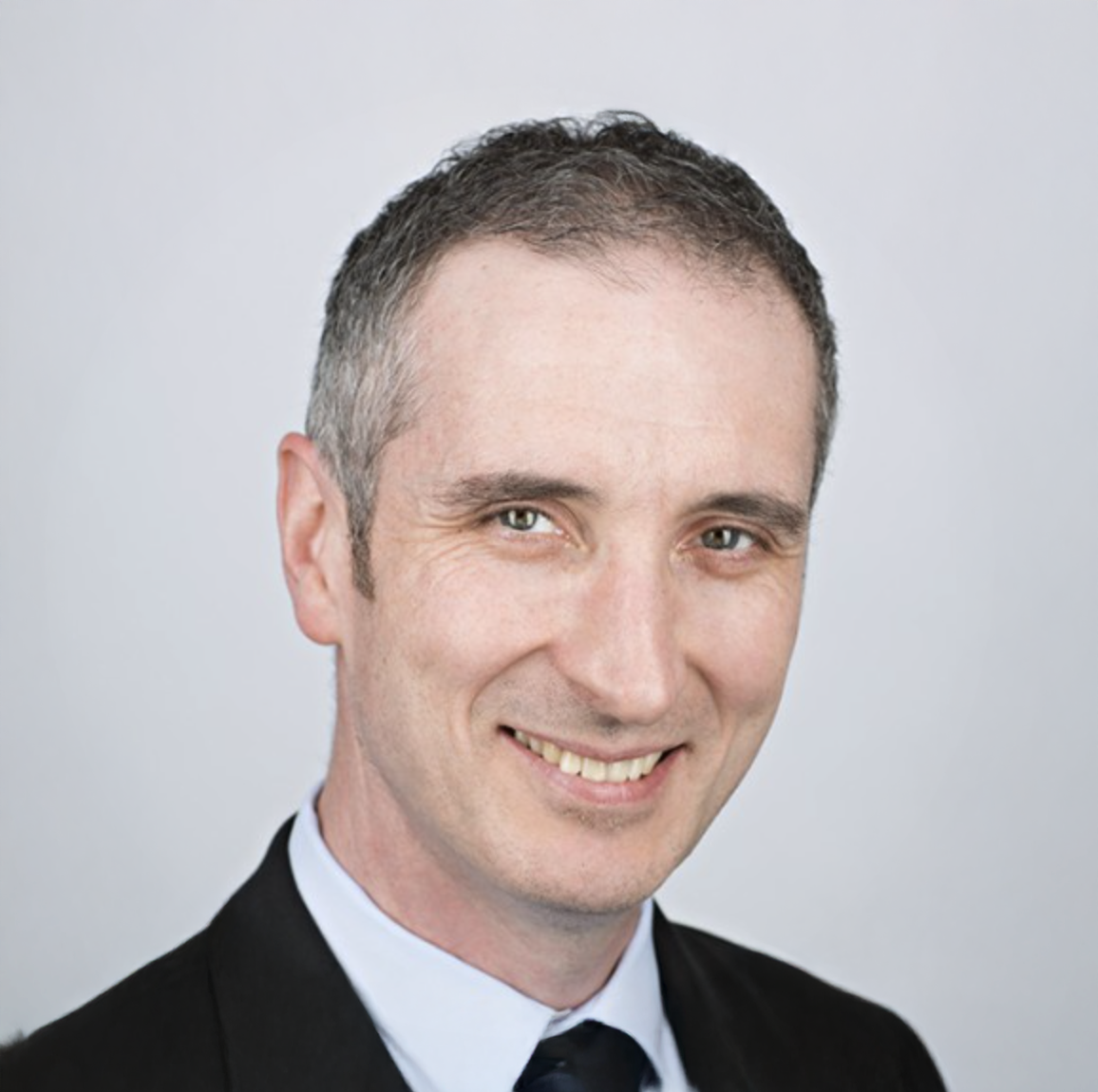 Professional headshot of a smiling middle-aged man with short gray hair wearing a dark suit and tie.