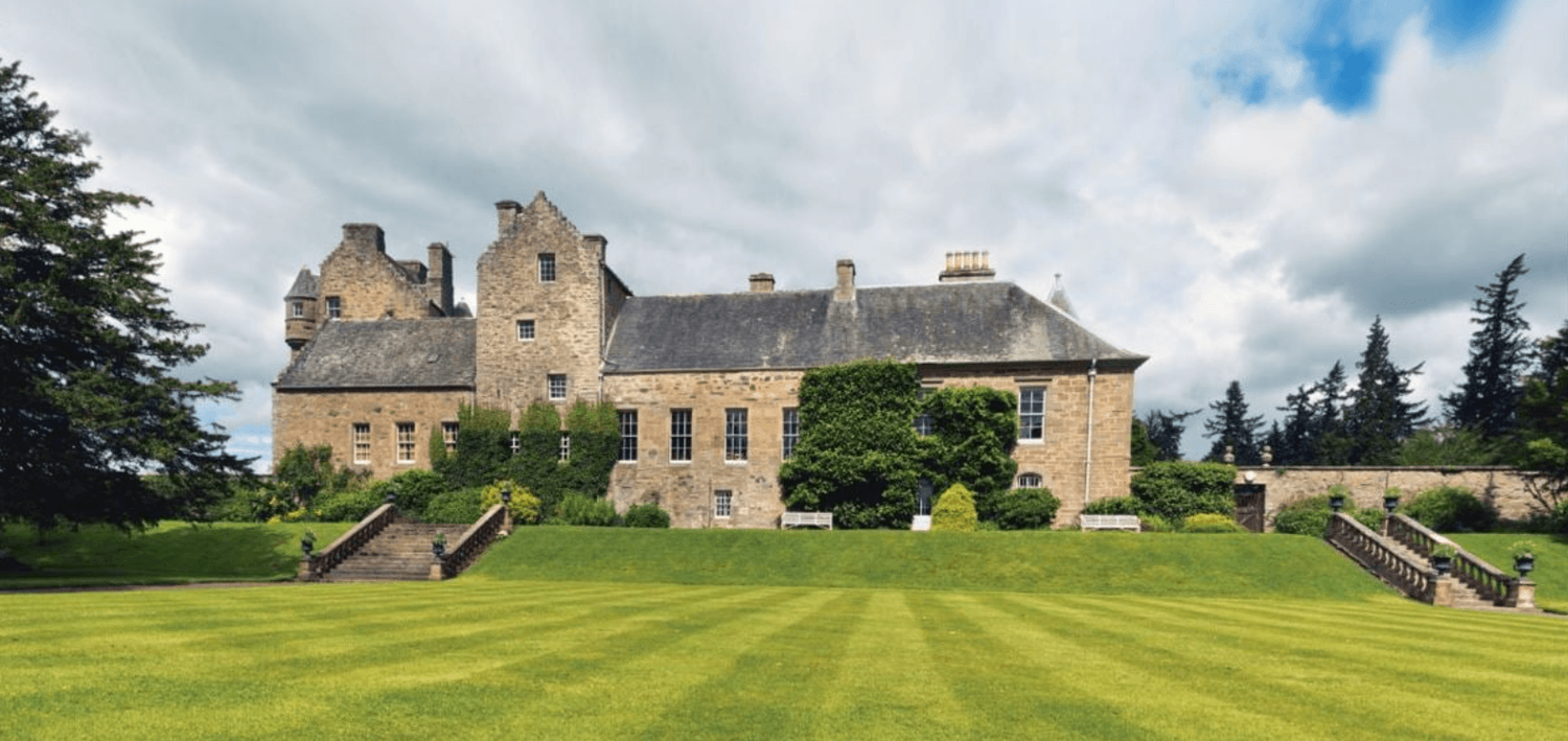 Historic Scottish castle with manicured green lawn, stone facade, and turrets under a cloudy sky.