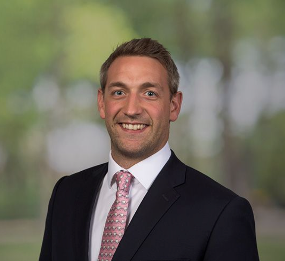 Professional headshot of a smiling businessman in a dark suit and pink tie outdoors.