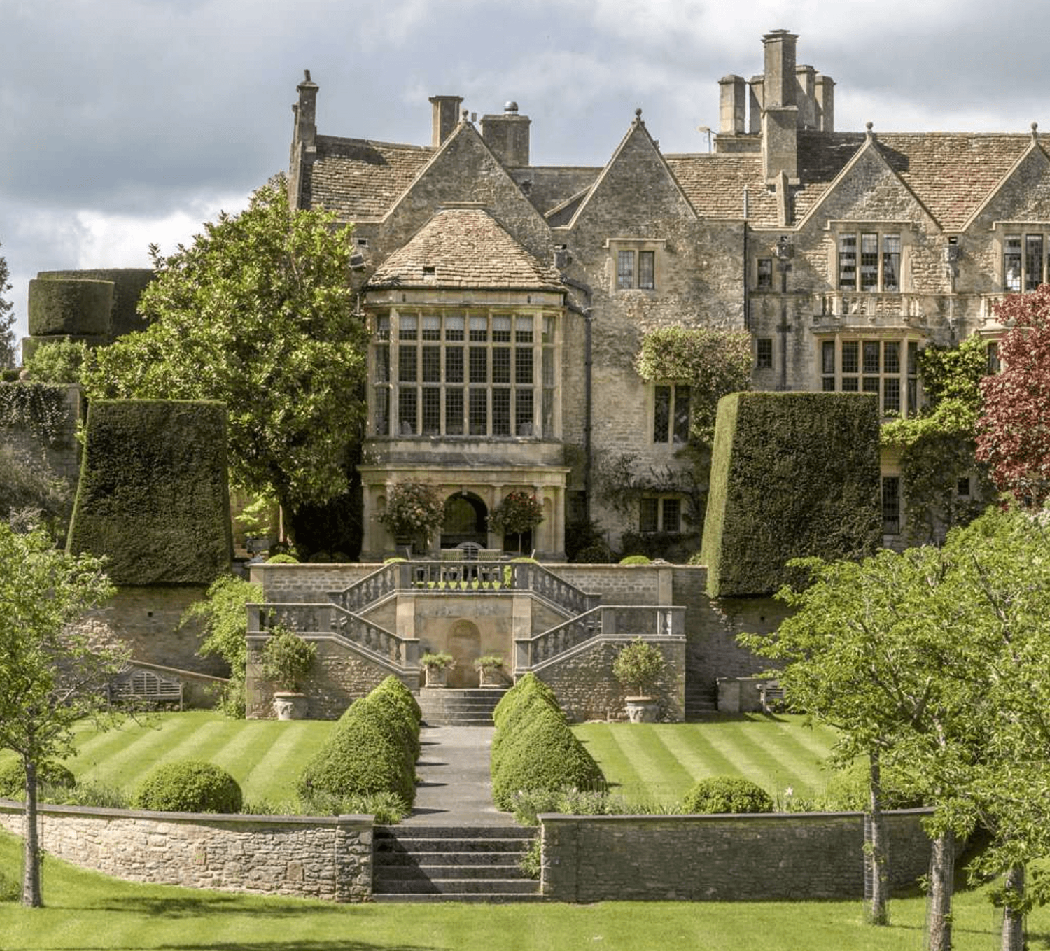 Grand English manor house with manicured topiary gardens, stone staircases, and lush green lawns under a cloudy sky.