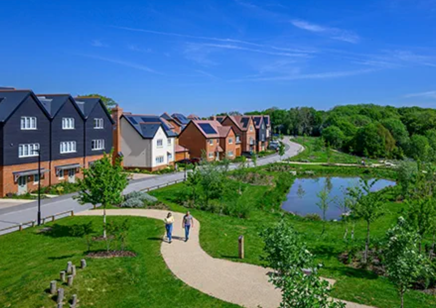 Modern housing development with solar panels, pond, and landscaped green spaces under a bright blue sky.
