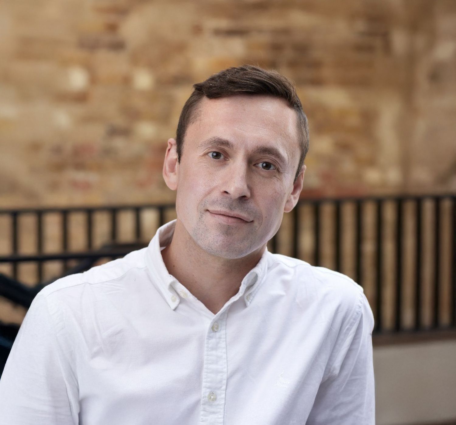 Professional headshot of a young man in a white shirt smiling, posed against a rustic brick wall background.
