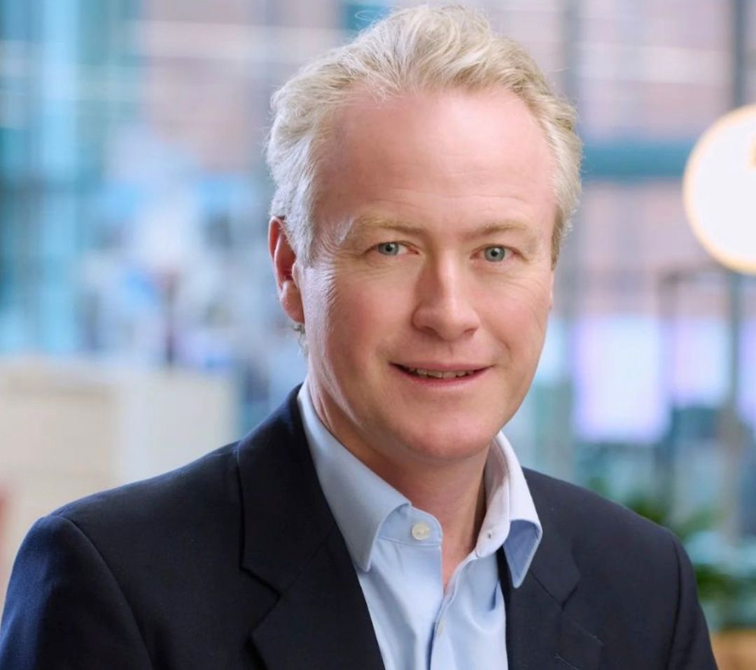 Professional headshot of a middle-aged man with silver hair wearing a dark blazer and light blue shirt in a modern office.