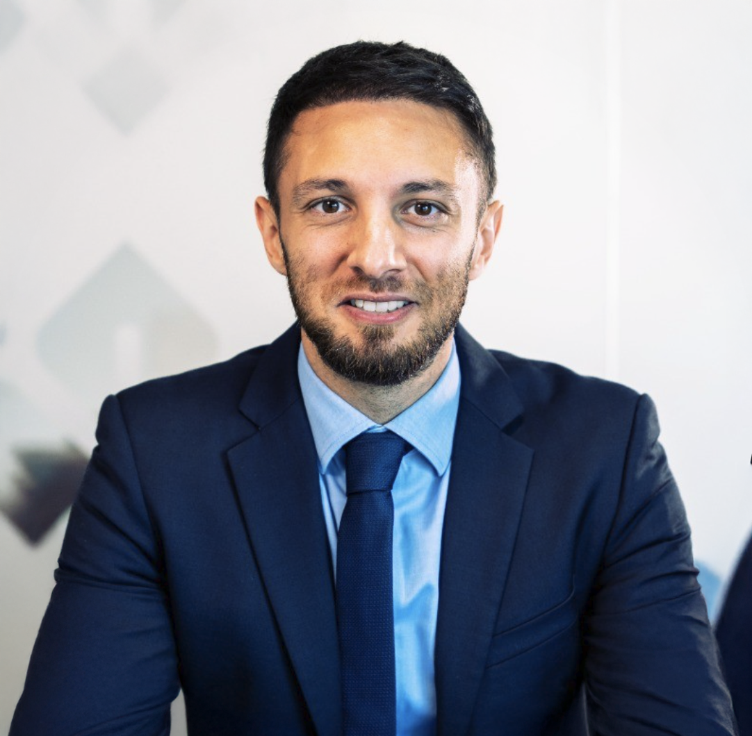 Professional businessman in navy suit and blue tie smiling confidently in a modern office setting.