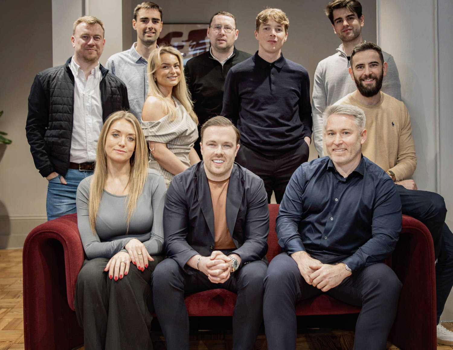 Group of eleven professionals posing for a team photo, seated and standing in front of a red sofa in a modern office.