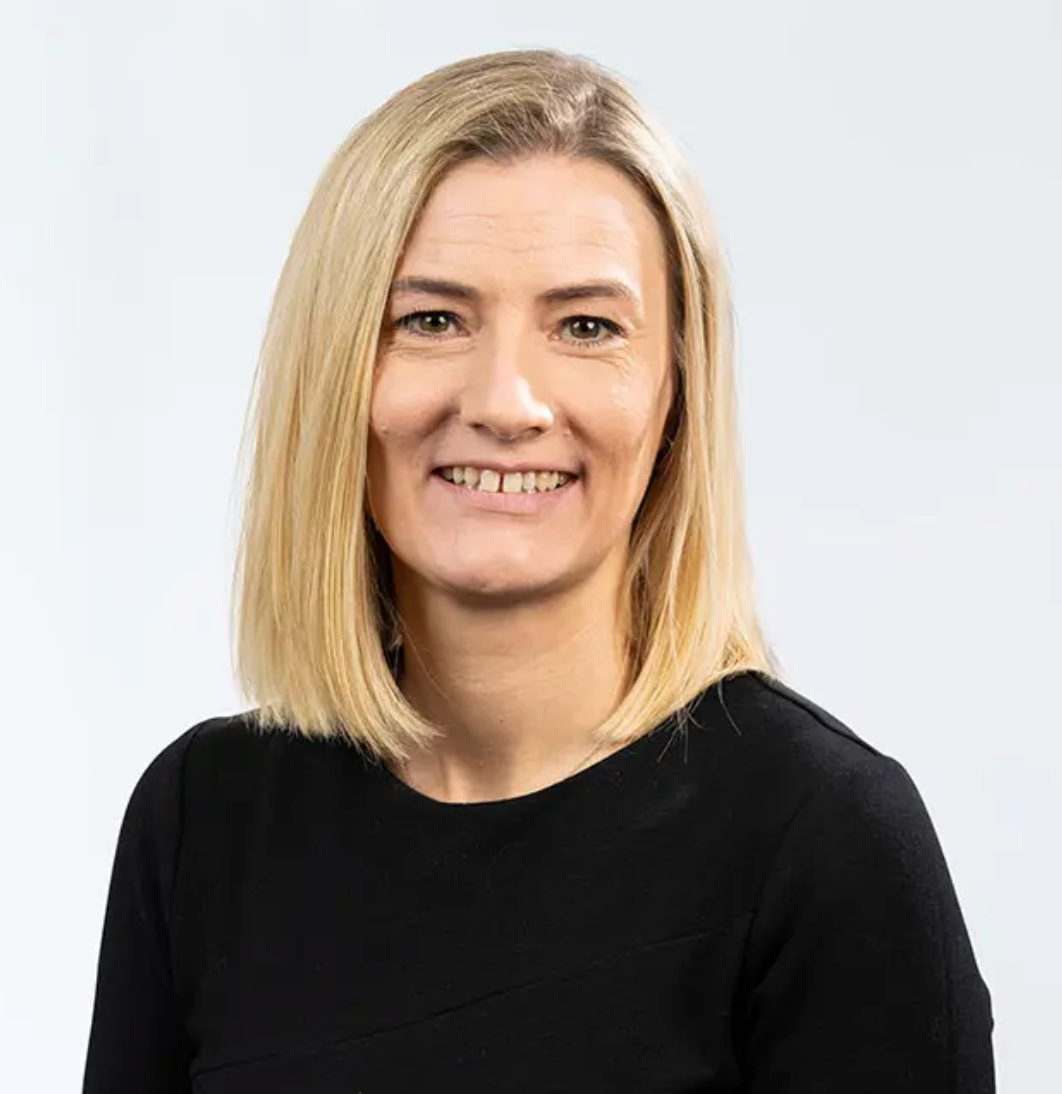 Professional headshot of a smiling blonde woman wearing a black top against a light gray background.