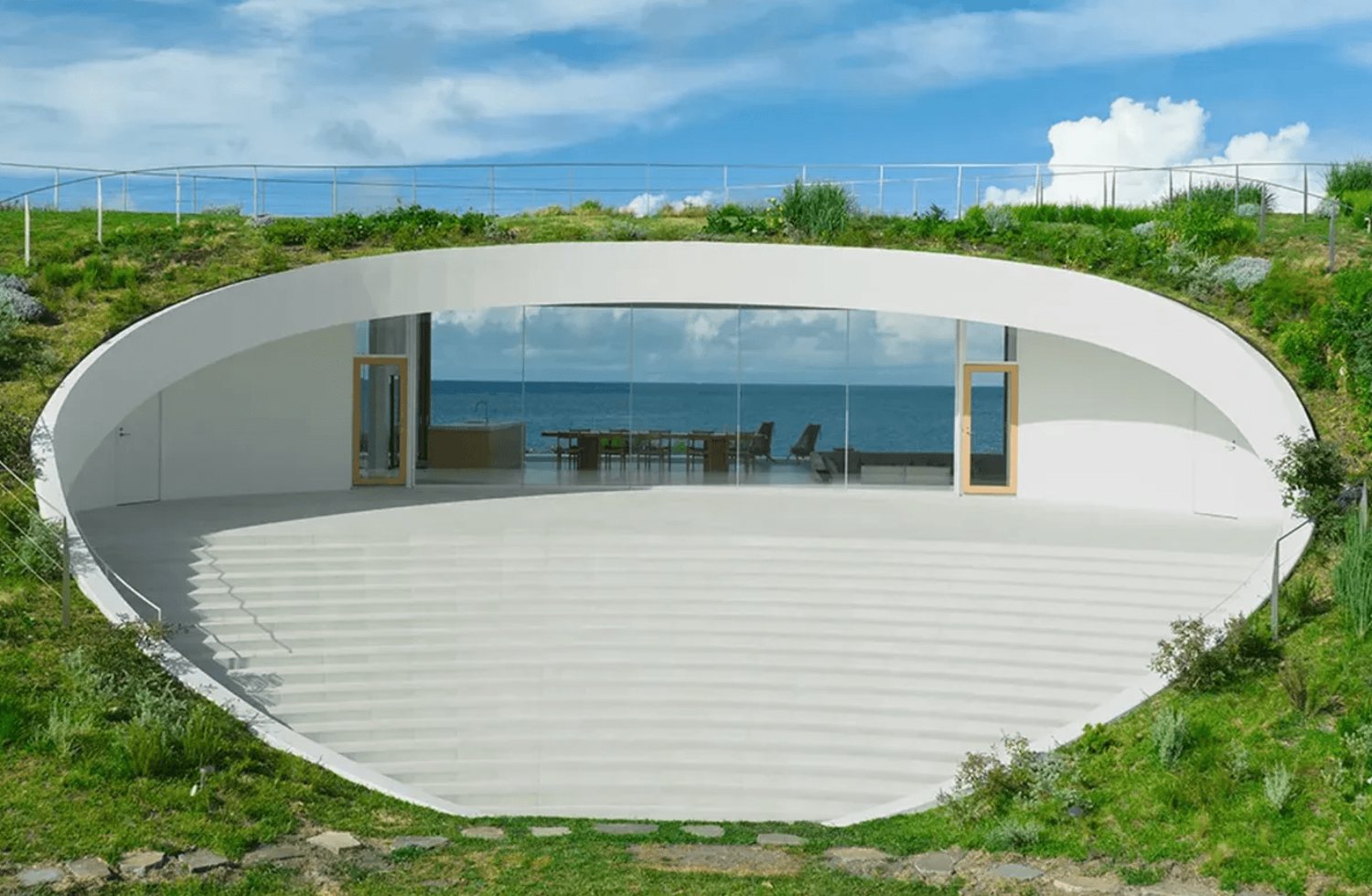 Modern oval-shaped white concrete structure with glass facade overlooking the ocean, set in lush green hillside landscape.