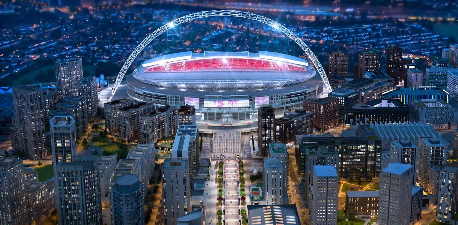 Aerial night view of Wembley Stadium illuminated in London, surrounded by city buildings and streets.