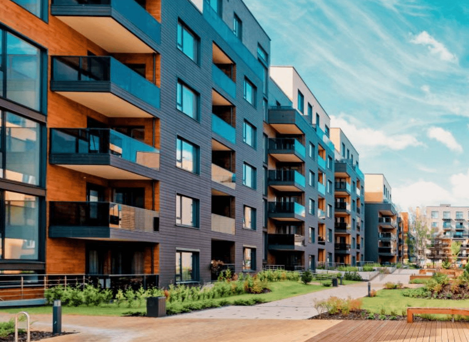 Modern apartment complex with blue balconies, wood accents, and landscaped courtyard under a bright blue sky.