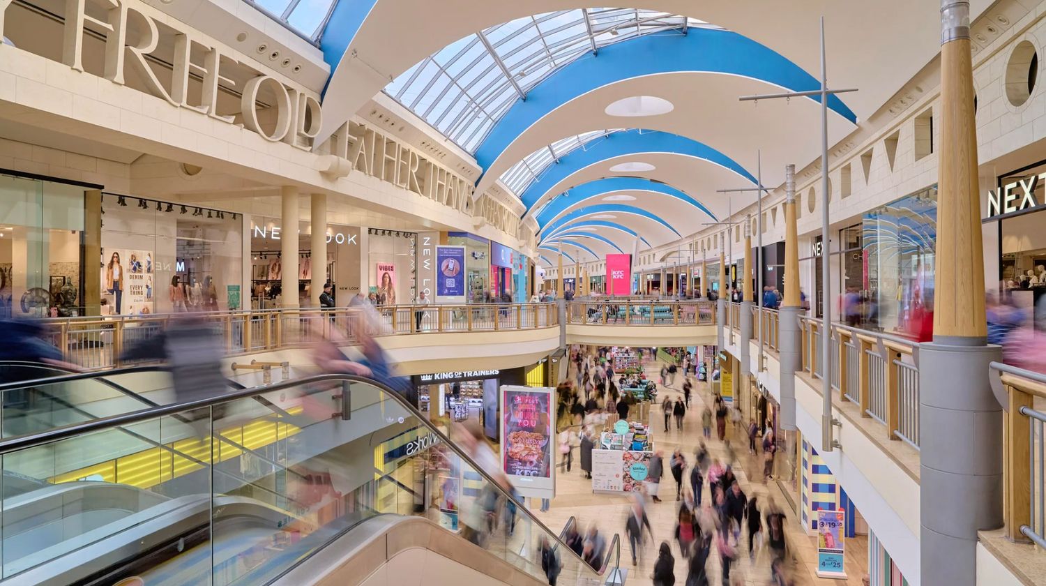 Busy multi-level shopping mall interior with escalators, blue arched ceiling, and crowds of shoppers browsing retail stores.