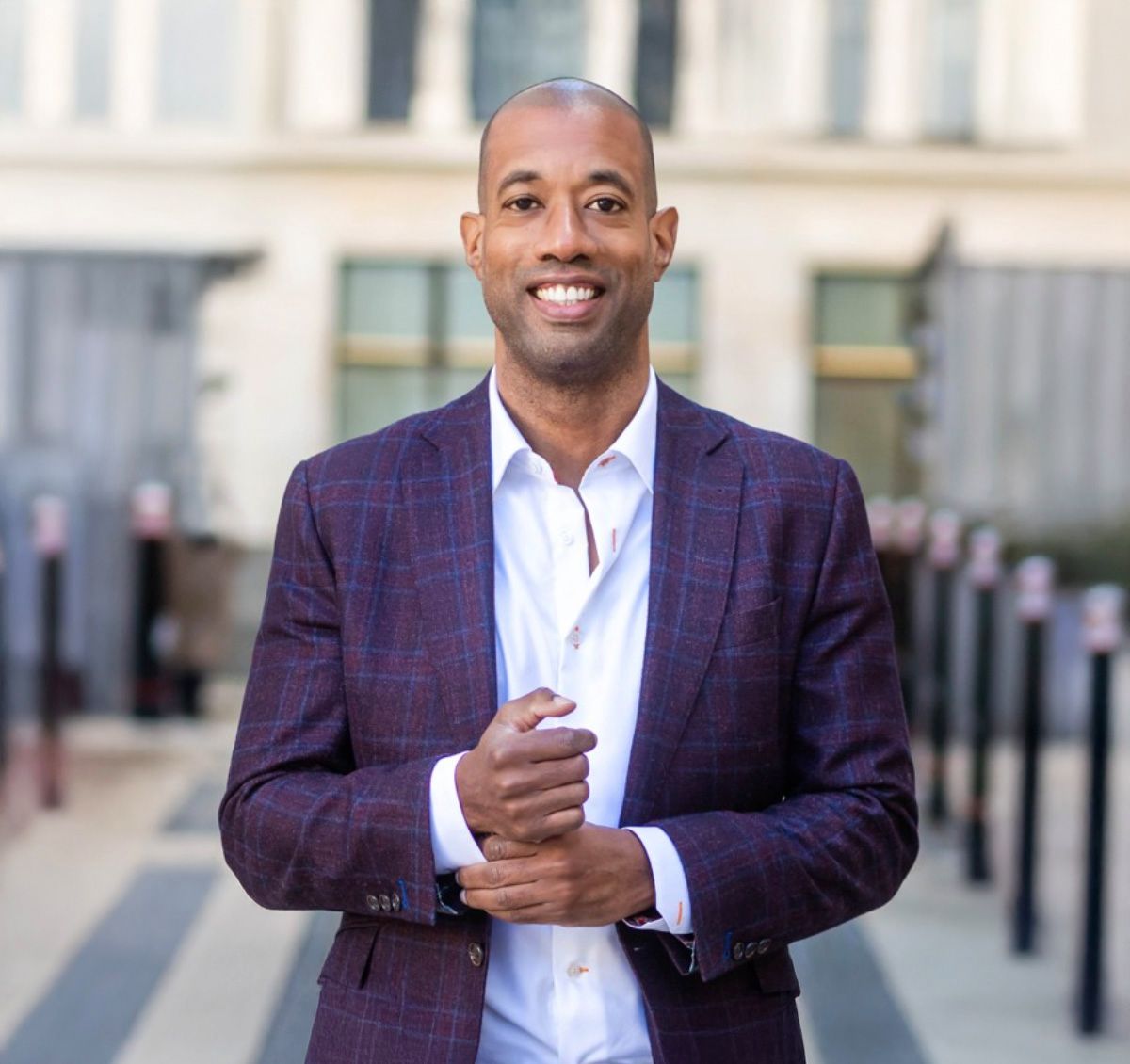 Smiling man in purple plaid blazer and white shirt poses confidently outdoors near a modern building.