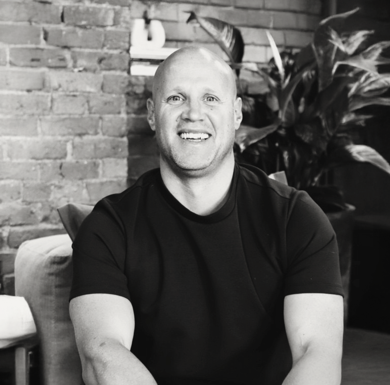Smiling bald man in black t-shirt seated in modern cafe with exposed brick wall and tropical plants.