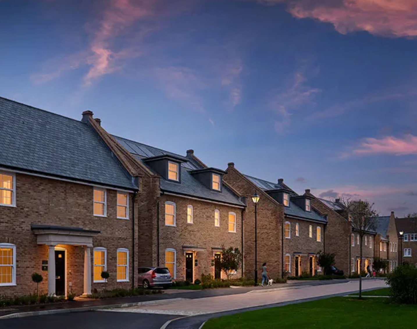 Row of modern brick townhouses at dusk with warm glowing windows under a vibrant pink and blue sunset sky.