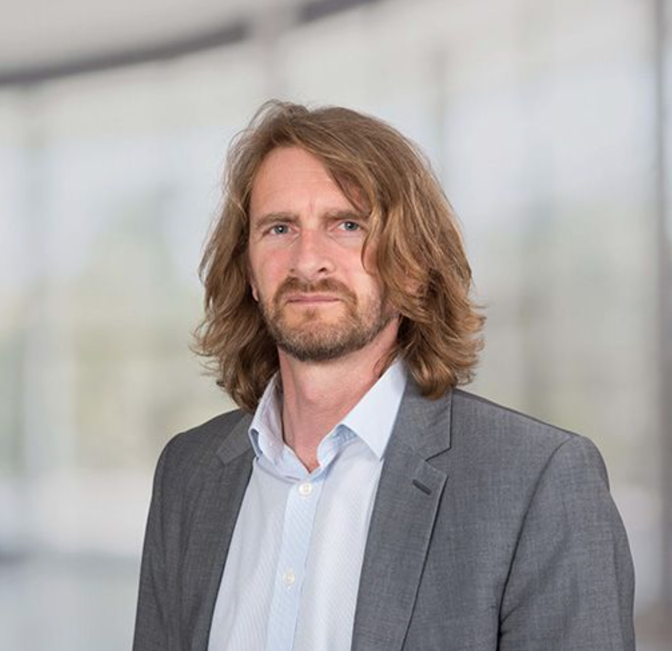 Professional man with shoulder-length hair and beard wearing a gray blazer in a modern office setting.