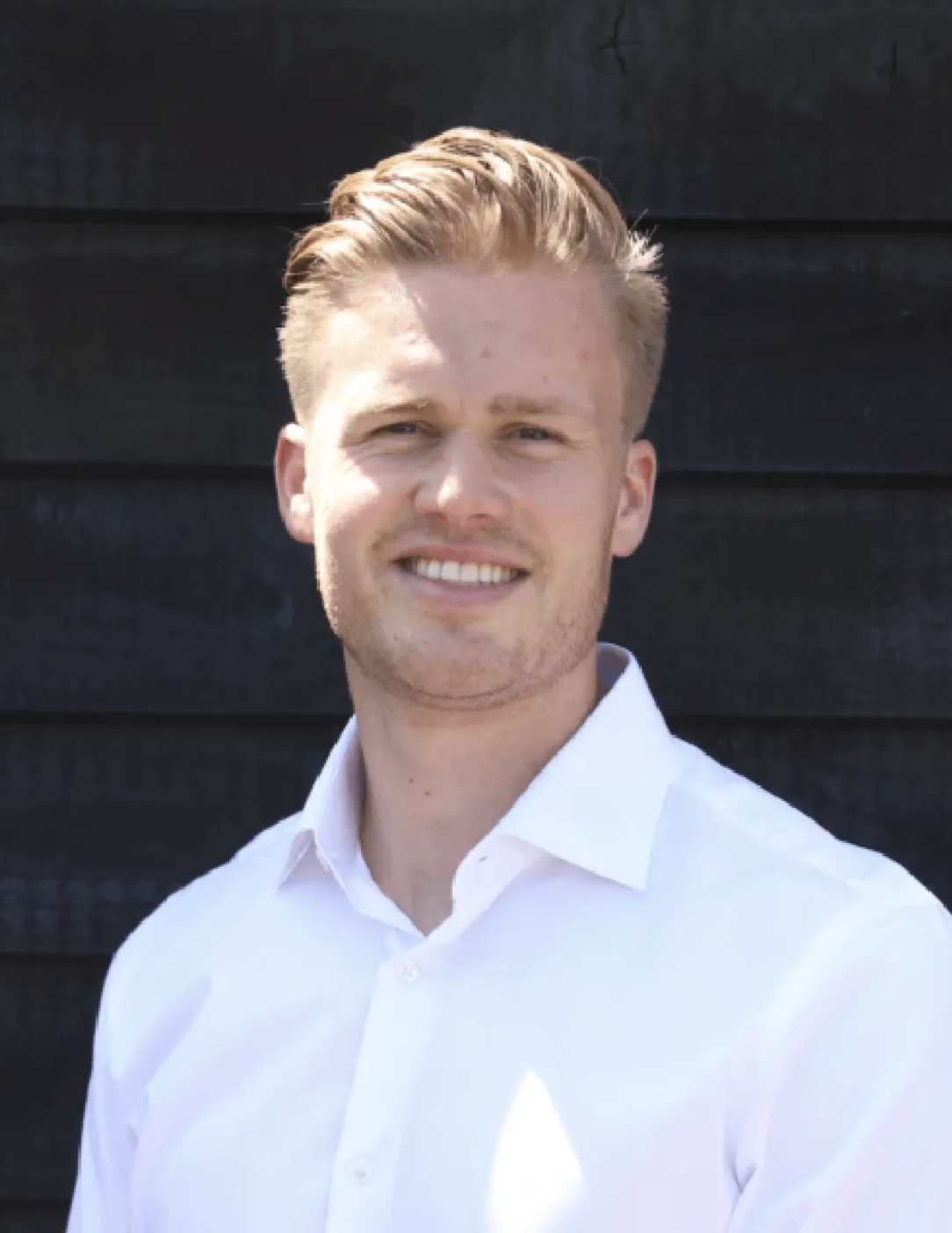 Young professional man with blonde hair smiling confidently in a white shirt against a dark wooden background.