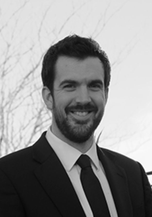 Professional headshot of a smiling bearded man in a black suit and tie outdoors.