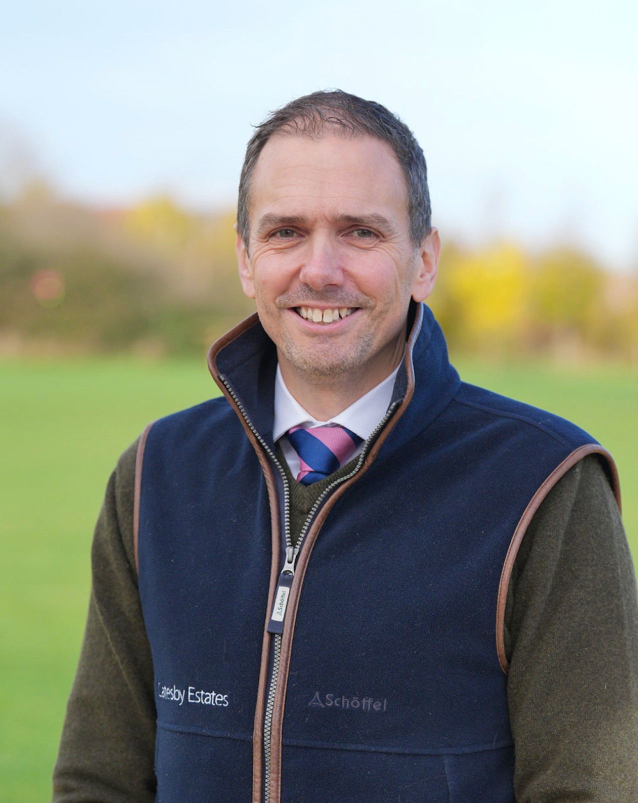 Man in navy Schoffel gilet and tweed jacket smiling outdoors in a green field on a bright autumn day.