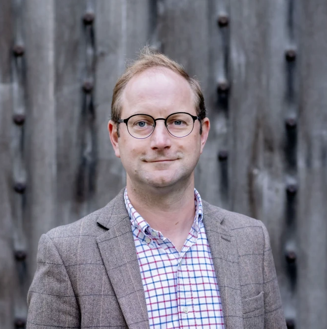 Professional man with glasses in a gray blazer and plaid shirt smiling outdoors against a wooden backdrop.