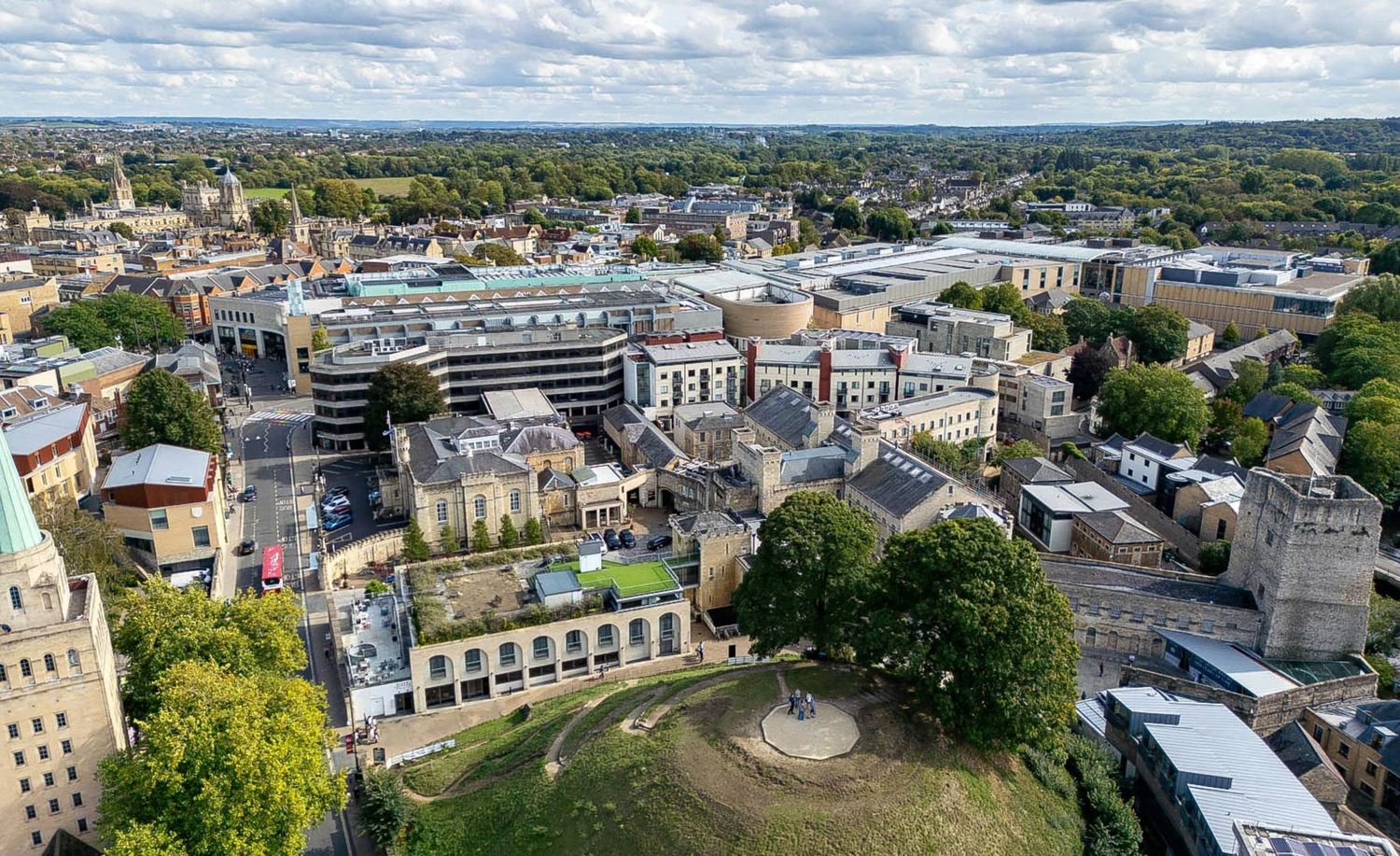 Aerial view of Oxford city center showing historic buildings, green trees, and modern structures under a clear sky.